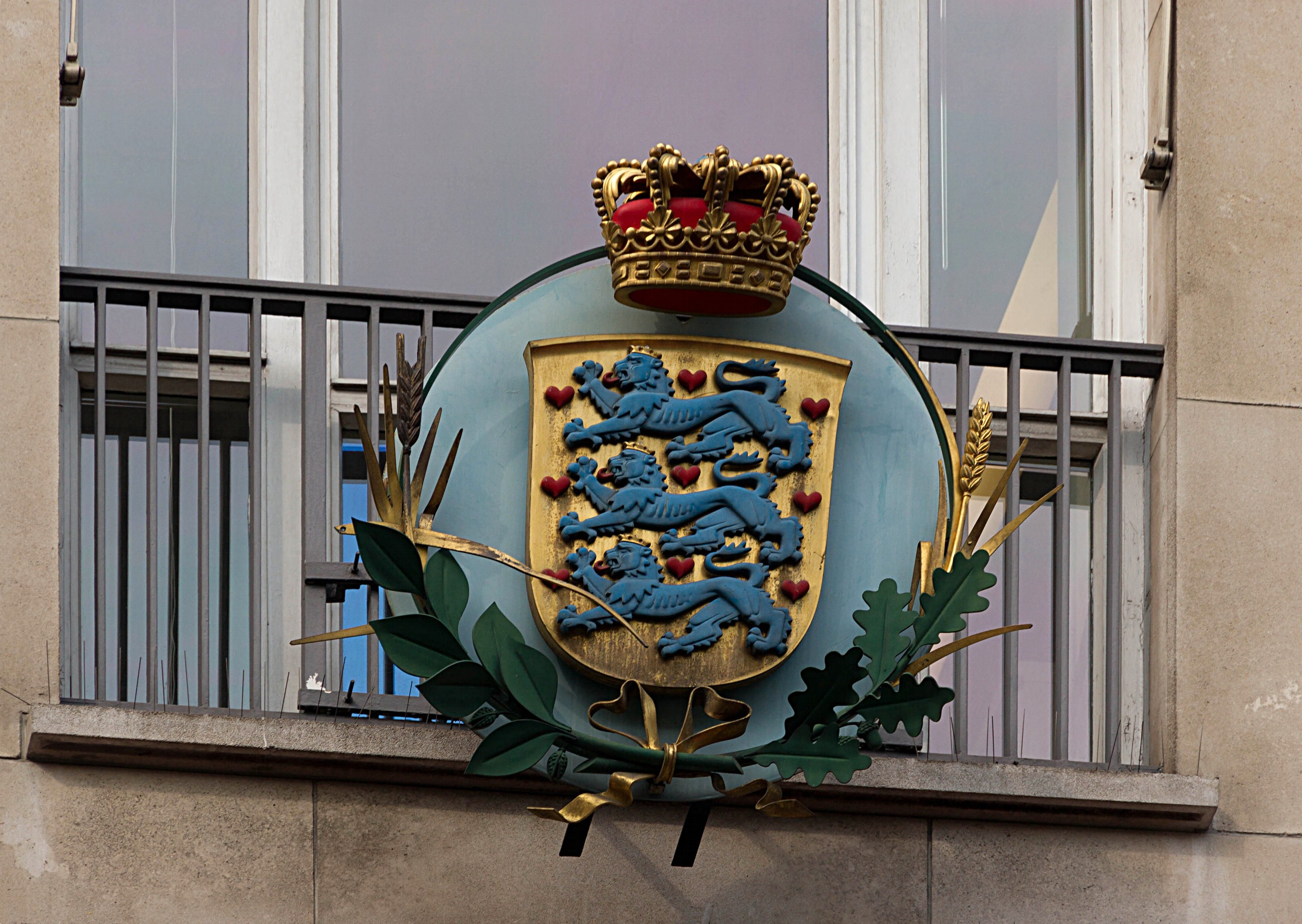 Wappen der britischen Königsfamilie auf der Außenseite eines Gebäudes dargestellt, begleitet von einem Logo mit Krone und Blättern, mit Fenstern und Gittern im Hintergrund.