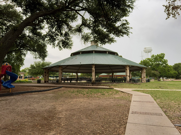 Ein Park mit einem Pavillon in der Mitte, umgeben von Bäumen, Bänken und einer Rutsche, mit einem Weg, der dorthin führt, und einem Wassertank im Hintergrund unter einem klaren blauen Himmel.