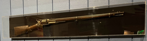 U.S. Springfield Model 1873 Gewehr in einer Glasvitrine mit Messingbeschlag, Holzschaft, Metalllauf und Messingabzugbügel.