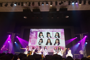 Eine Gruppe von Menschen performs auf einer hell beleuchteten Bühne mit Trägern, während ein Sänger ein Mikrofon hält und ein Musiker ein Instrument spielt, während das Publikum unten die Vorstellung mit Kameras aufzeichnet und ein großer Bildschirm Bilder der K-Pop-Gruppe BK48 anzeigt.