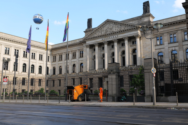 Großes modernes Gebäude mit vielen Fenstern, als Bundestag in Berlin, Deutschland identifiziert, umgeben von Straßeninfrastruktur, Fußgängern, Flaggen und Bäumen unter einem bewölkten Himmel.