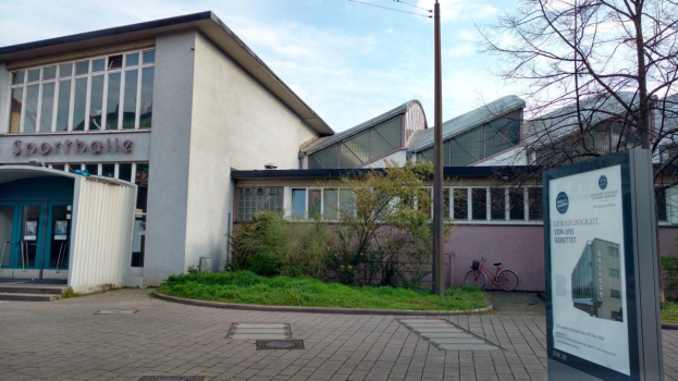 Ein großes Gebäude mit einem Schild davor, umgeben von Bäumen, Pflanzen, Gras, Fahrrädern und einem Pfahl, mit einem bewölkten Himmel im Hintergrund, als Sportballe bekannt, soll 2020 eröffnet werden.