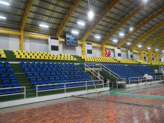 Großer Hallensportplatz mit Holzboden, blauen und gelben Sitzplätzen, Geländern, Treppen zum Sitzbereich, wandmontierten Bildschirmen, Deckenbeleuchtung und einigen wenigen Menschen.