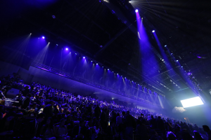 Eine große Menschenmenge sitzt in einer Konzerthalle und schaut auf eine Bühne mit Deckenleuchten und einem Bildschirm auf der rechten Seite.