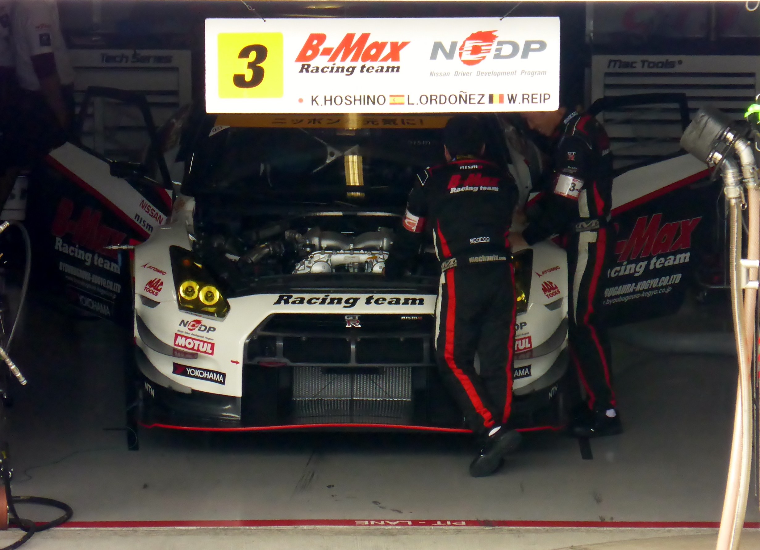 Ein Nissan GT-R Nismo GT3 Rennwagen in einer Garage mit zwei Personen, die daran arbeiten, Rohre und Gegenstände auf der rechten Seite und Tafeln mit Text und Zahlen im Hintergrund.