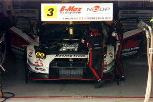 Ein Nissan GT-R Nismo GT3 Rennwagen in einer Garage mit zwei Personen, die daran arbeiten, Rohre und Gegenstände auf der rechten Seite und Tafeln mit Text und Zahlen im Hintergrund.