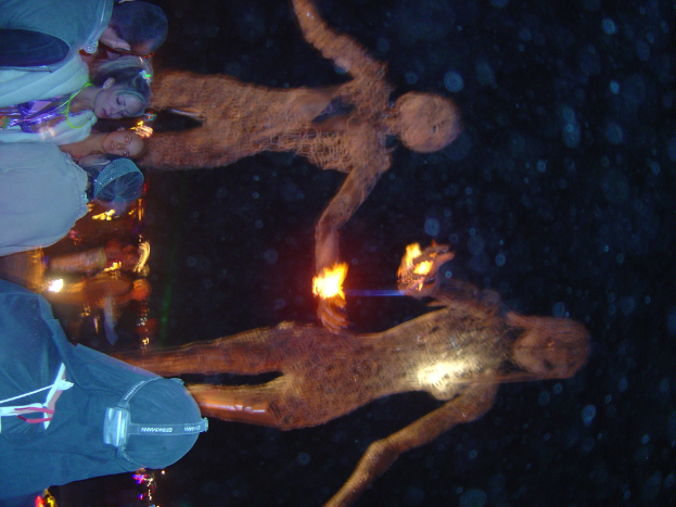 Eine Gruppe von Menschen steht auf der linken Seite des Bildes, hinter ihnen brennen Statuen und im Hintergrund leuchten Lichter.