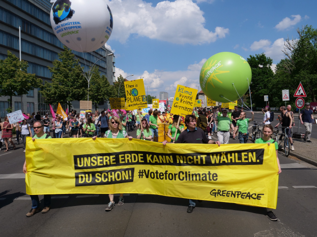 Eine Gruppe von Menschen, die eine Straße entlanggehen und ein gelbes "Wähle das Klima"-Plakat halten, mit Ballons, Schildern, Fahrrädern, Bäumen, Laternenmasten und Gebäuden im Hintergrund unter einem klaren blauen Himmel.