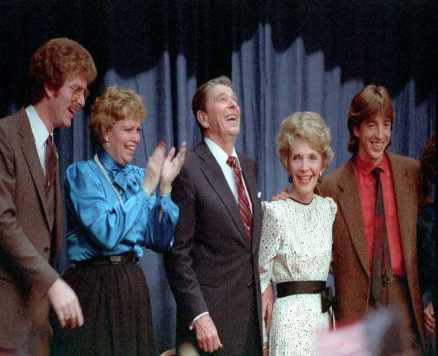 Group of people from the TV show 'The Brady Bunch' standing on a stage, smiling and clapping with a curtain in the background and objects at the bottom.