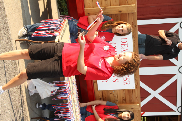 Eine Frau steht mit einer Medaille in der Hand, mit mehreren Medaillen auf einem Tisch, drei stehende Personen, eine Person hält ein Mikrofon und eine Tafel auf einem Holzzaun im Hintergrund.