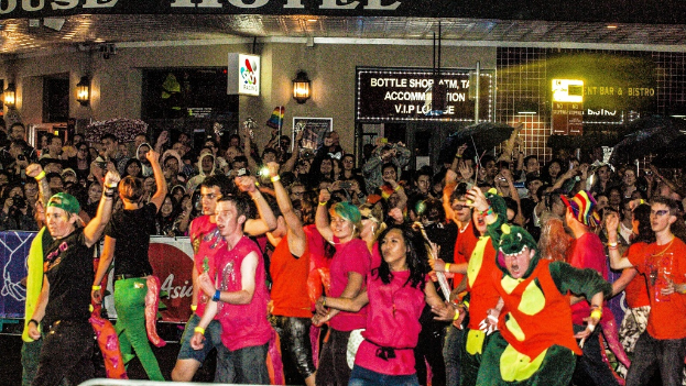 Eine Gruppe von Menschen, die tanzen und Spaß haben, vor einem Gebäude mit Schildern, Wandlampen, einem Grill und einem Banner auf der Straße dahinter.