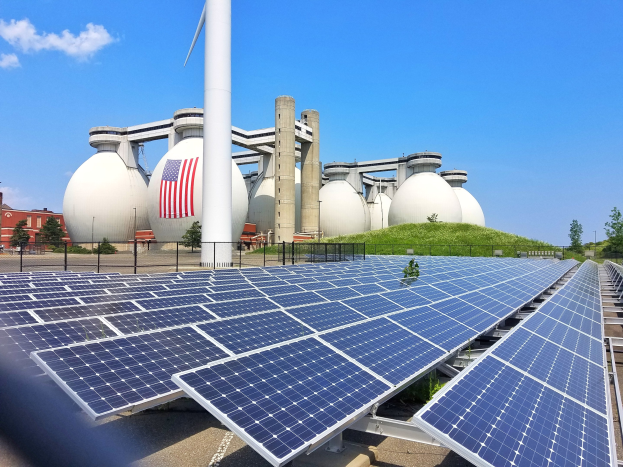 Solarpanels auf einem Gebäudedach mit einer Flagge, umgeben von einem Zaun, Grünfläche und anderen Strukturen, vor einem bewölkten Himmel.