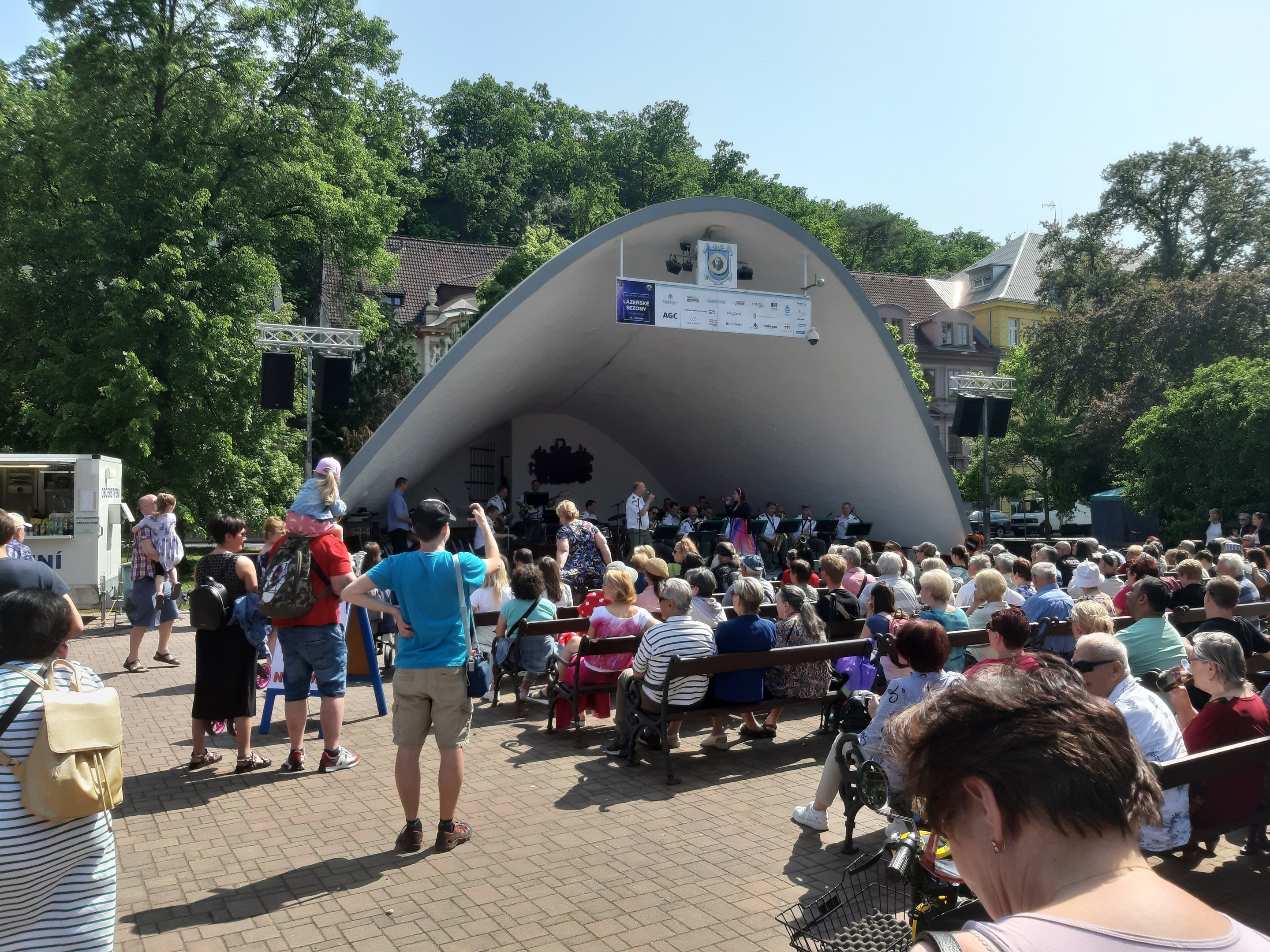 Menschen auf Bänken und Stehplätzen vor einer Bühne mit einem Fotografen, einem Stativ, einer Fahne, Pfosten, einem Metallrahmen, Bäumen, Gebäuden mit Fenstern und einem bewölkten Himmel bei einem Sommerkonzert im Park.