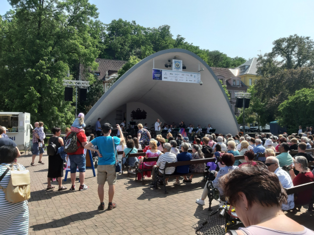Menschen auf Bänken und Stehplätzen vor einer Bühne mit einem Fotografen, einem Stativ, einer Fahne, Pfosten, einem Metallrahmen, Bäumen, Gebäuden mit Fenstern und einem bewölkten Himmel bei einem Sommerkonzert im Park.