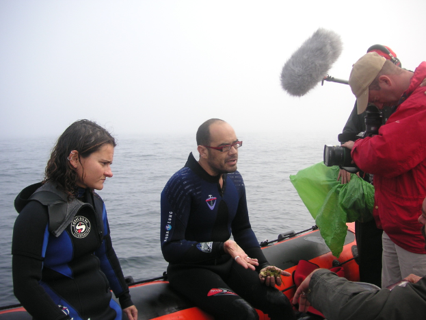 Eine Gruppe von Menschen sitzt auf einem Boot, mit einer Frau in Jacke und Hose links, einem Mann mit Brille, T-Shirt und Hose neben ihr, einem Mann mit Kamera rechts und einer weiteren Person mit Mütze, T-Shirt und Kopfhörern, die ein Mikro hält, mit Taschen in der rechten unteren Ecke, dem Ozean im Hintergrund und dem Himmel mit Wolken darüber.