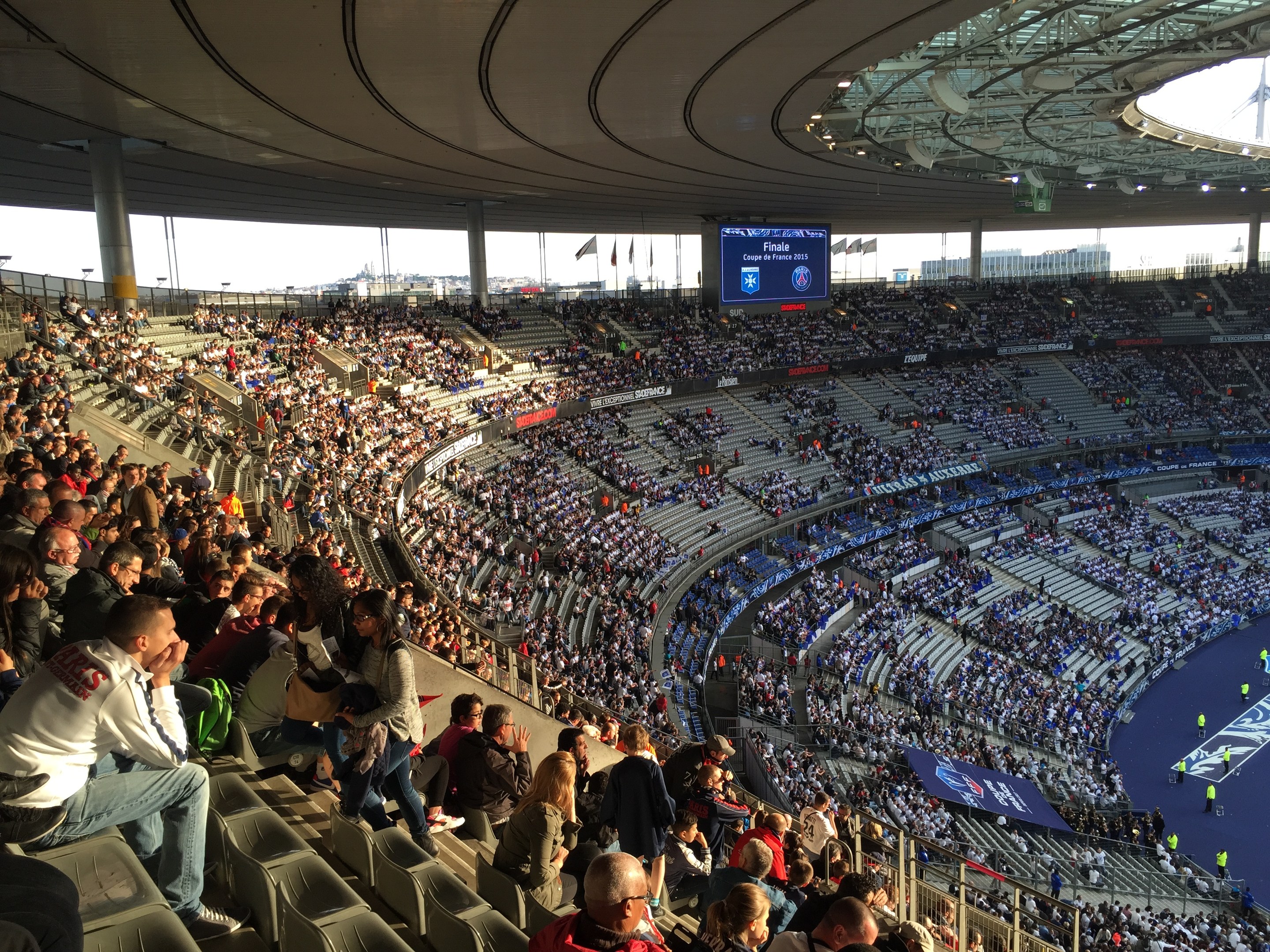 Große Zuschauermenge im Allianz Arena Stadion bei einem Fußballspiel, mit einer Bühne, Fahnen und einem Bildschirm im Hintergrund.