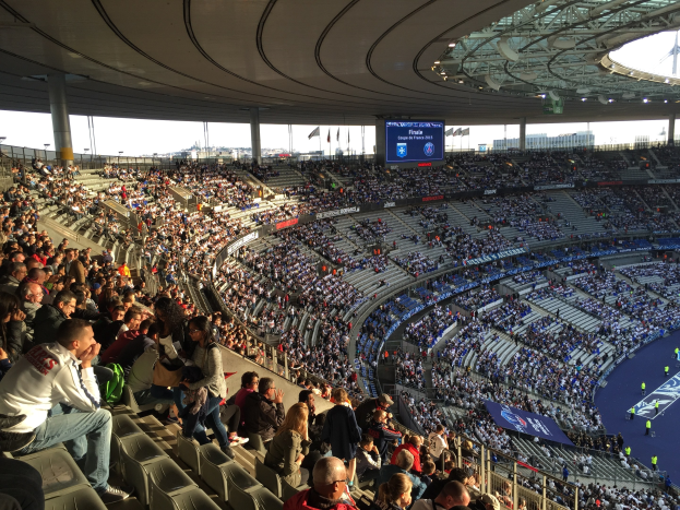 Große Zuschauermenge im Allianz Arena Stadion bei einem Fußballspiel, mit einer Bühne, Fahnen und einem Bildschirm im Hintergrund.