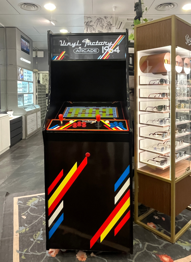 Ein Geschäft mit einem vintage Arcade-Automaten in der Mitte, einem Gestell mit Gläsern auf der rechten Seite und verschiedenen Gegenständen wie Schränken und einem Fernseher im Hintergrund, alles unter Deckenlampen, die eine retrohafte Atmosphäre schaffen.