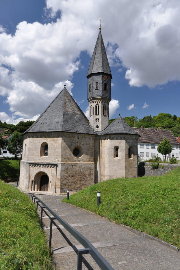 Alte steinerne Kirche mit einem Turm und einem Glockenturm in Lauterbrunnen, Deutschland, umgeben von einer grünen Wiese, Gebäuden, Bäumen und einem bewölkten Himmel.