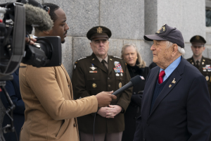 Mann im Anzug und Kappe, der in ein Mikrofon spricht, während ein Reporter ein Objekt hält und ein Videograf im Hintergrund aufnimmt.