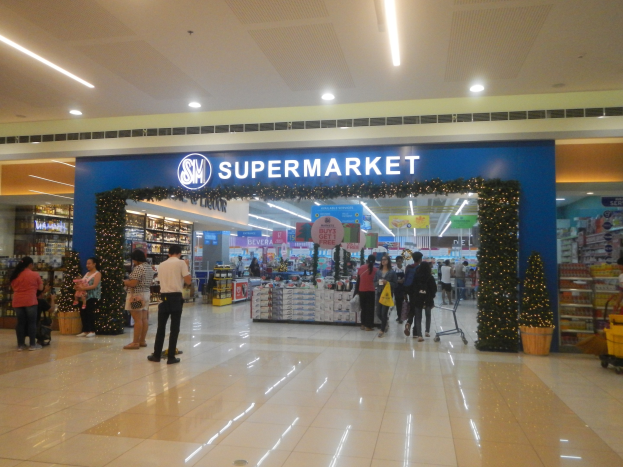 Eine Gruppe von Menschen, die vor einem Supermarkt stehen, einige halten Einkaufstäsche, mit einem Einkaufswagen, Topfpflanze, Regalen, Schildern und Deckenbeleuchtung.