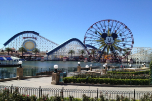 Disneylands California Adventure mit einem Riesenrad und Achterbahn, umgeben von Grünflächen und Laternenmasten, mit Wasser im Vordergrund unter einem blauen Himmel.