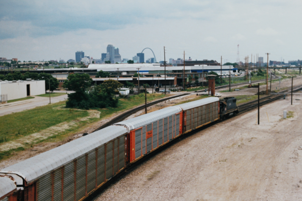 Ein Güterzug fährt auf Schienen neben einer Stadt, mit Gebäuden, Bäumen, Gras, Strommasten, Kabeln und einem bewölkten Himmel im Hintergrund.